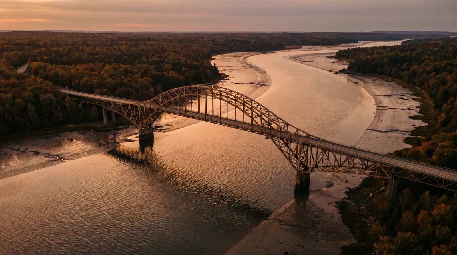 Golden-hour aerial of a steel arch highway bridge crossing a tidal river between wooded New England shorelines