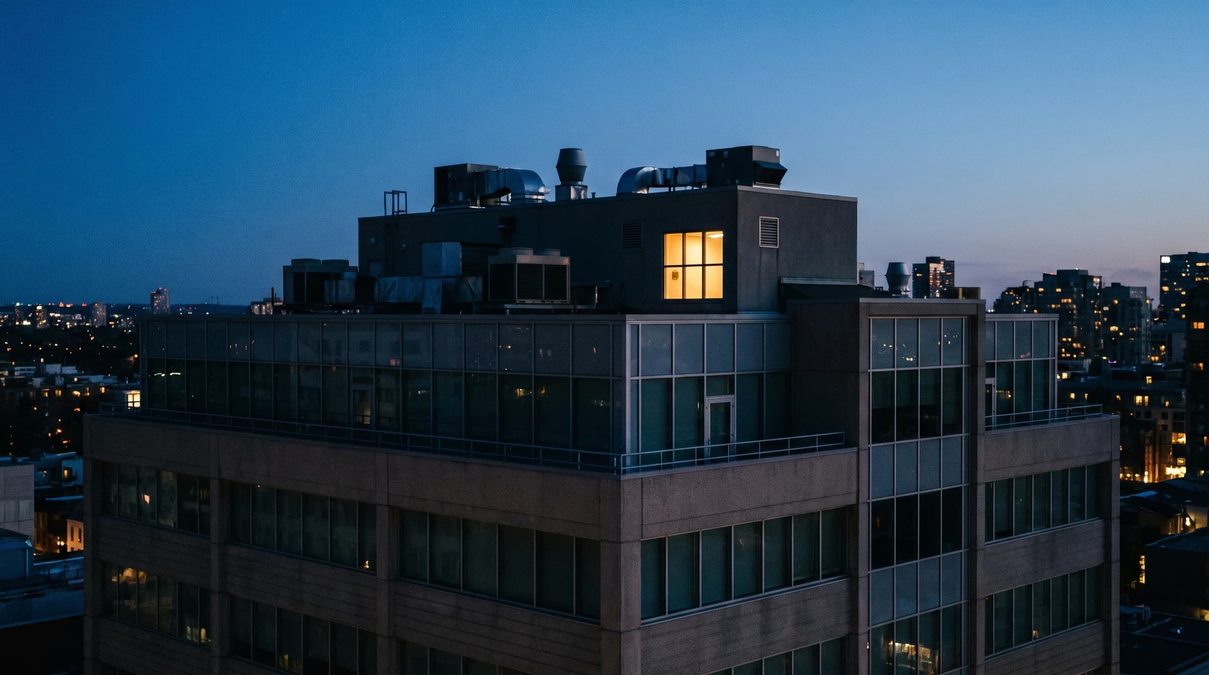 Blue-hour commercial building with rooftop mechanical silhouettes and a lit penthouse plant room