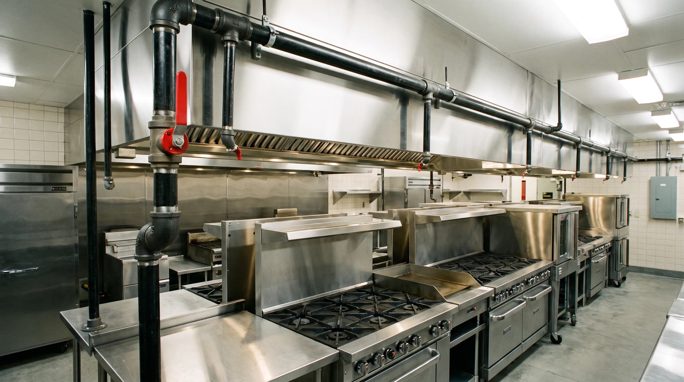 New black iron gas drops with red emergency shutoff feeding stainless ranges in a bright commercial kitchen